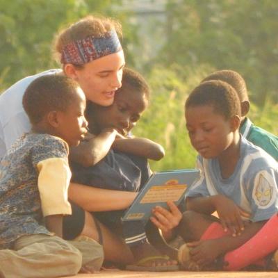 Projects Abroad Childcare volunteer reads to three children outside a kindergarten to develop their literacy skills in Ghana. A volunteer with children in Ghana reads a book to a group of young students outdoors at her Childcare Project.